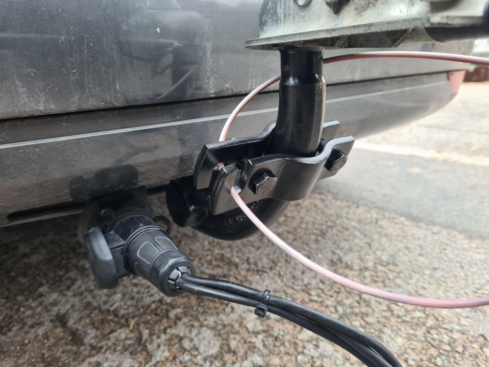 Hecksto&szlig;stange eines grauen Autos mit Anh&auml;ngerkupplung, schwarzer Steckdose und zwei Kabeln, kiesiger Untergrund