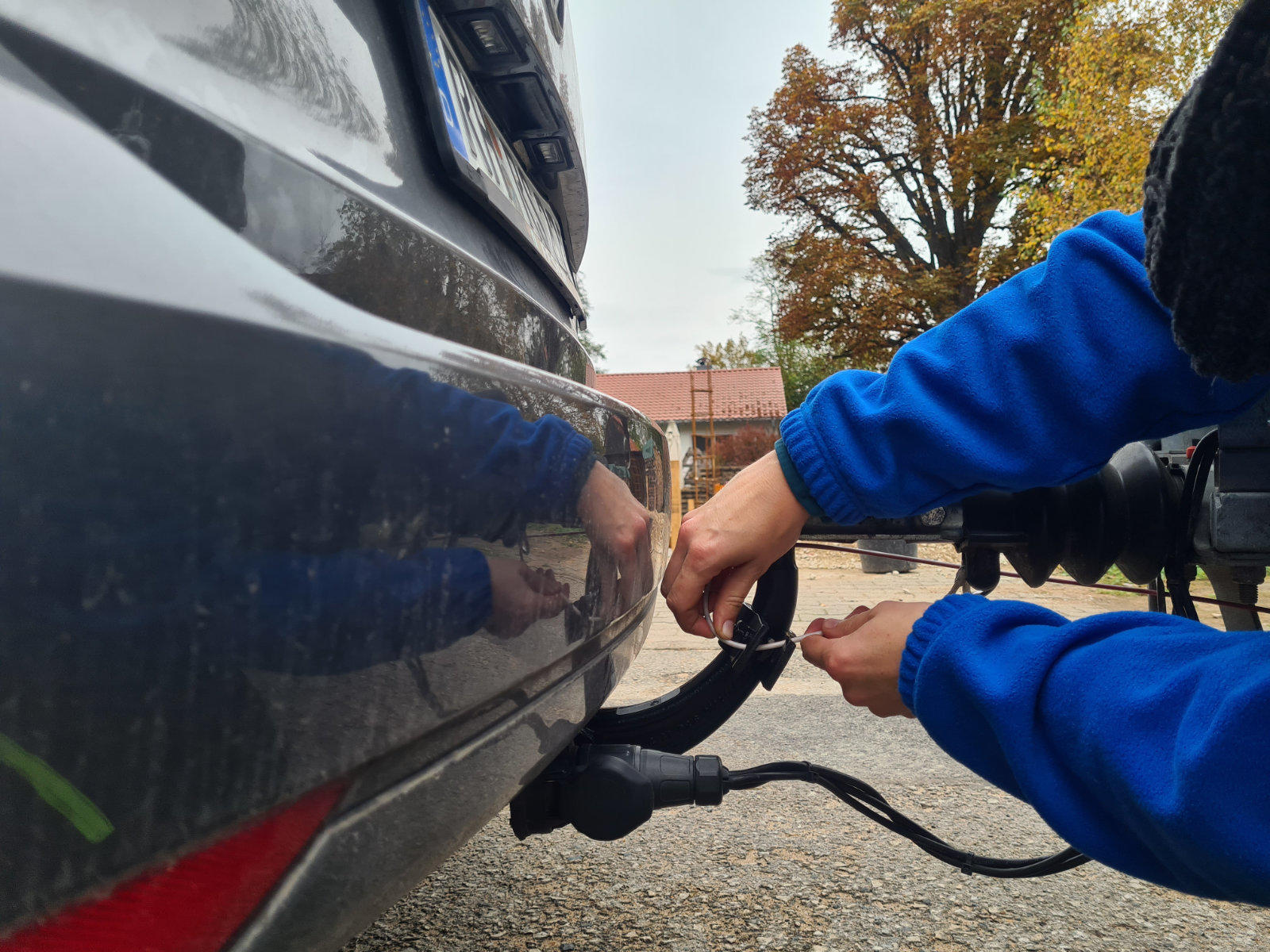 H&auml;nde in blauem Jacken&auml;rmel stecken einen Stecker in die Anh&auml;ngerkupplung eines Autos; Hecksto&szlig;stange, Pflaster und B&auml;ume im Hintergrund