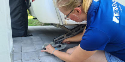 Person kniet auf gepflastertem Boden und dreht mit einer Kurbel am St&uuml;tzrad unter einem wei&szlig;en Anh&auml;nger; ein Kabel liegt daneben