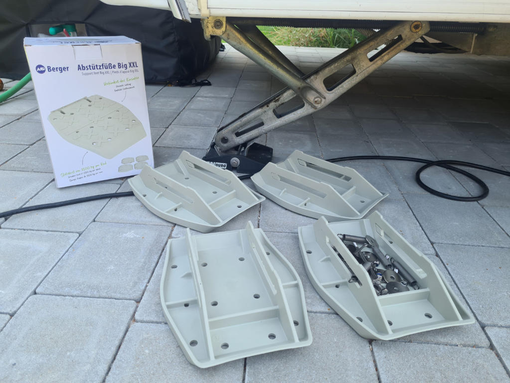 Mehrere helle Kunststoffplatten und ein offenes Beh&auml;ltnis mit Schrauben liegen auf grauem Pflaster neben einer ausgeklappten Metallst&uuml;tze unter einem Fahrzeug; daneben eine Kartonverpackung und ein schwarzer Beutel