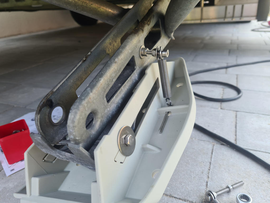 Nahaufnahme eines metallischen Scherenwagenhebers, befestigt an einer hellgrauen Kunststoffhalterung; Schrauben und Muttern auf gefliestem Boden