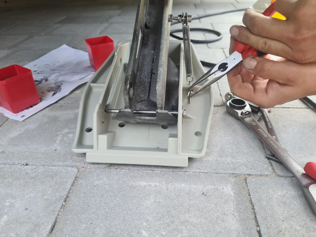 H&auml;nde mit roter Zange halten eine Schraube an einem Metallwinkel auf einer grauen Kunststoffplatte auf Pflaster; Schraubenschl&uuml;ssel, rote Kunststoffkeile und Papier im Hintergrund