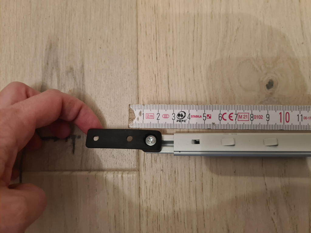 Linke Hand h&auml;lt schwarzes Metallst&uuml;ck mit Bolzen; rechts eine wei&szlig;e Metallschiene; auf der Schiene liegt ein Lineal mit Zentimeter-Einteilung; alles auf hellem Holzuntergrund