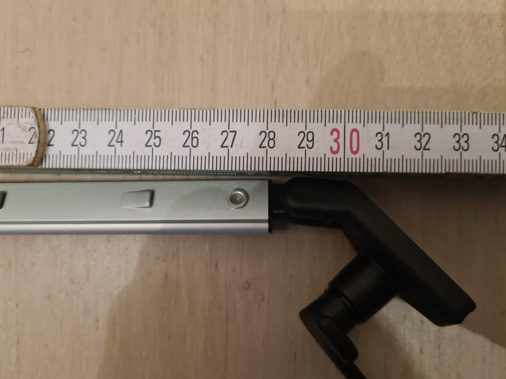 Ma&szlig;band mit sichtbarer Skala bis 34 cm und roter &sbquo;30&lsquo;, eine silberne Metallf&uuml;hrung darunter und ein schwarzer L-f&ouml;rmiger Kunststoffanschluss auf hellem Holzuntergrund