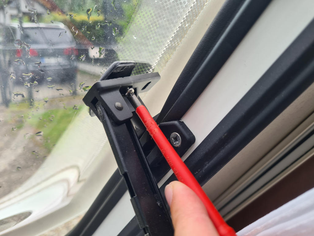 Hand h&auml;lt roten Schraubendreher, Spitze auf Schraube in schwarzer Fensterhalterung am Rahmen; regennasse Fensterscheibe und unscharfes Fahrzeug im Hintergrund