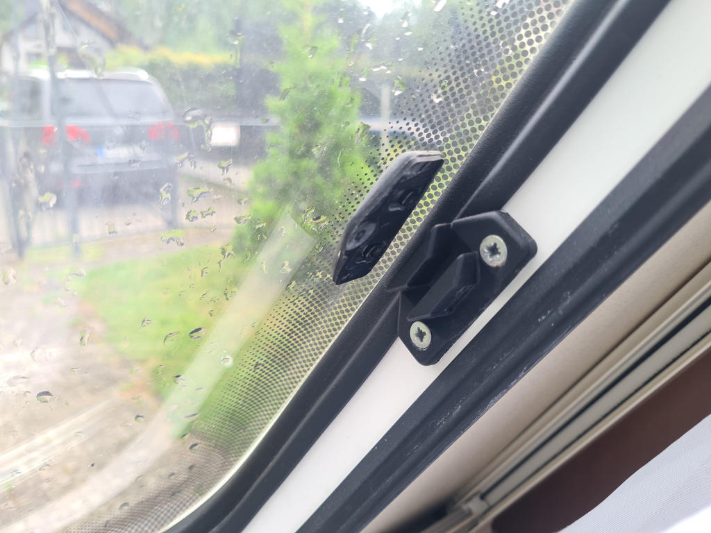 Nahaufnahme eines Fensterverschlusses am Fenster mit Regentropfen auf der Scheibe; unscharfes Fahrzeug und Pflanzen drau&szlig;en