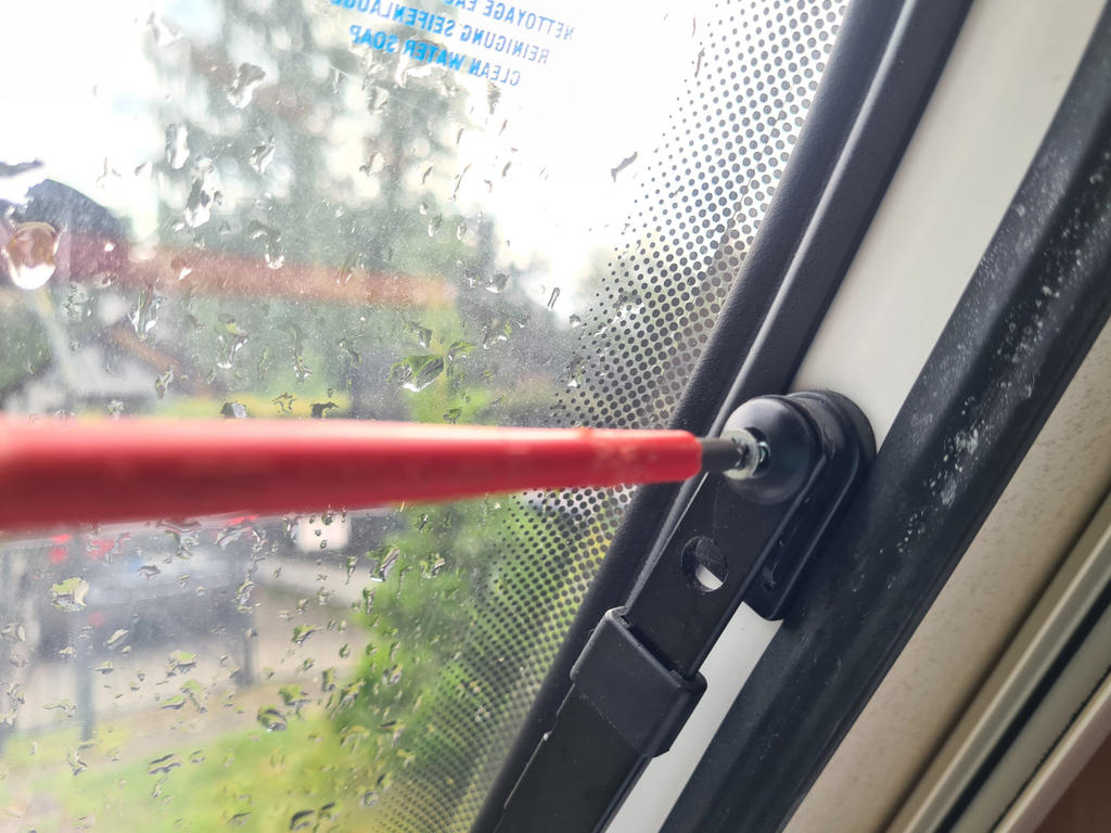 Nahaufnahme eines Fensters mit Regentropfen, schwarzes Befestigungsst&uuml;ck am Rand, roter Stab in einer Schraube, punktf&ouml;rmige Randbedruckung, unscharfer gr&uuml;ner Hintergrund