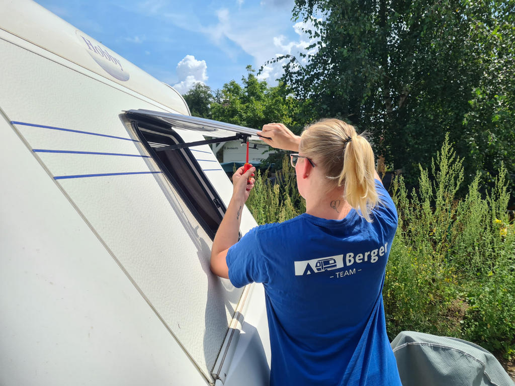 Person mit blondem Pferdeschwanz in blauem T-Shirt h&auml;lt einen Schraubenzieher an einem ge&ouml;ffneten Klappfenster eines Wohnwagens