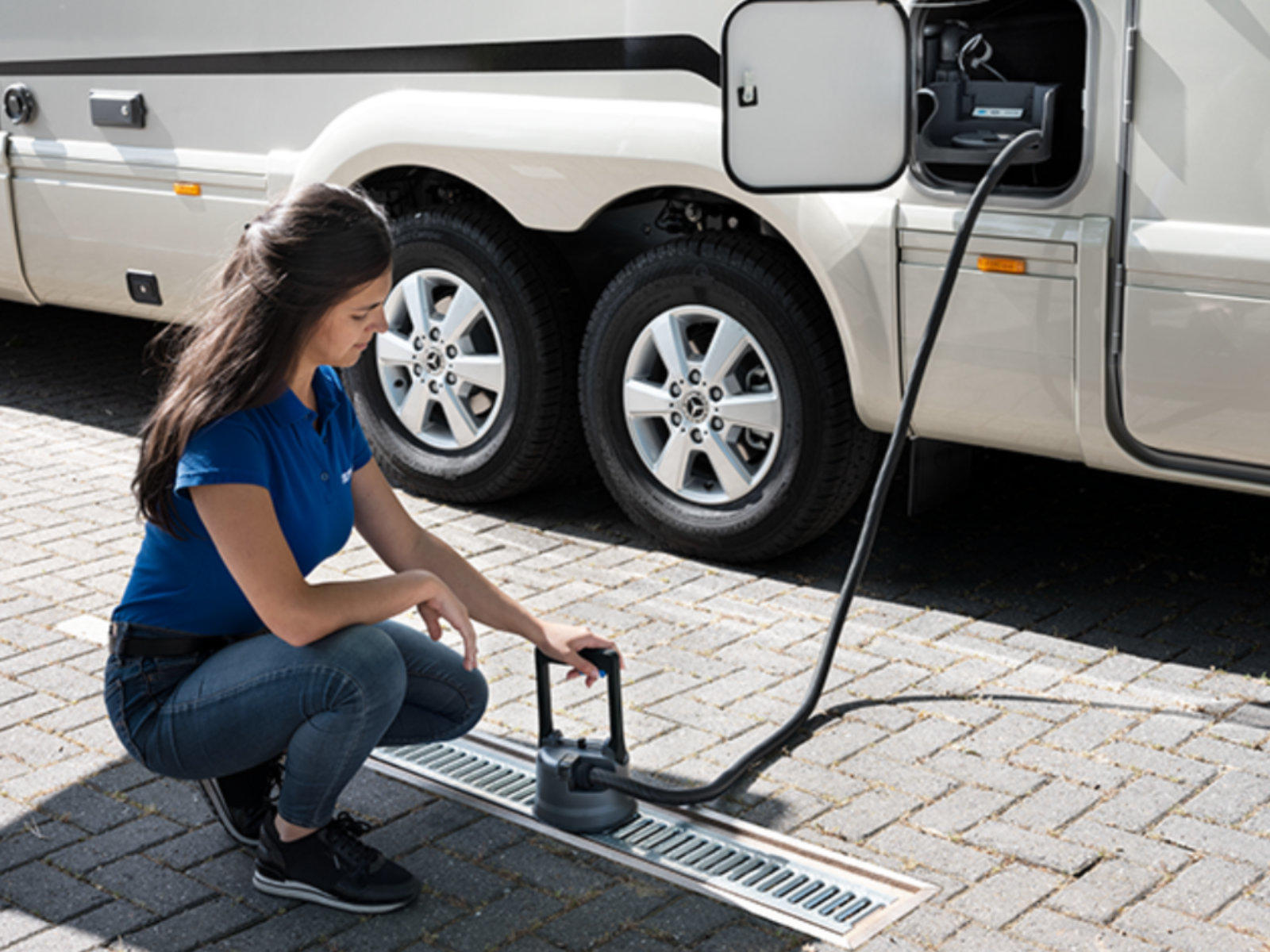 Eine Frau kniet auf gepflastertem Boden neben einem hellen Wohnmobil mit offener Klappe; ein schwarzes Kabel verl&auml;uft von der Klappe zum Boden und sie h&auml;lt ein rundes schwarzes Anschlussst&uuml;ck