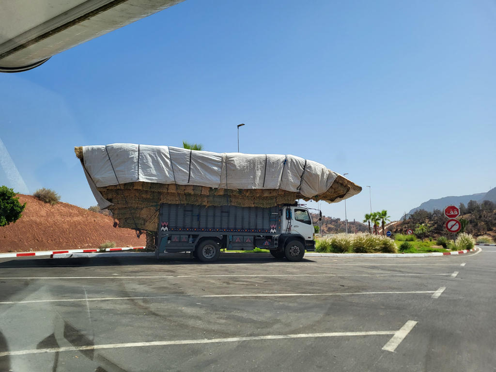 Lastwagen mit wei&szlig; abgedeckter Ladung, die seitlich &uuml;ber die Ladefl&auml;che hinausragt, auf einer Stra&szlig;e; blauer Himmel, Bordstein, Fahrbahnmarkierungen, Verkehrsschilder, niedrige Bepflanzung und H&uuml;gel im Hintergrund; Fensterrand eines anderen Fahrzeugs