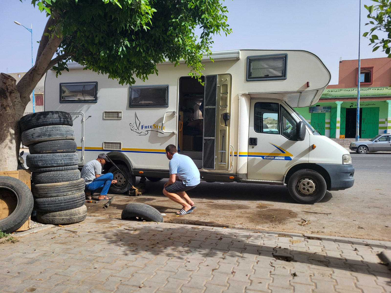 Wei&szlig;es Wohnmobil am Stra&szlig;enrand mit ge&ouml;ffneter T&uuml;r; zwei Personen neben dem vorderen Rad; mehrere Autoreifen liegen auf dem Boden neben einem Baum; Gehweg und H&auml;user im Hintergrund