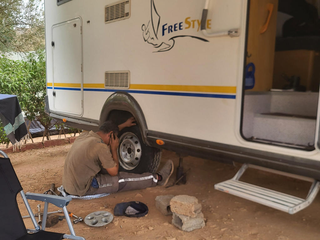 Person kniet neben dem Rad eines Wohnwagens; Radabdeckung, Radkappe und M&uuml;tze liegen auf dem Boden; ge&ouml;ffnete T&uuml;r und Trittstufe am Wohnwagen; Campingst&uuml;hle und B&auml;ume im Hintergrund