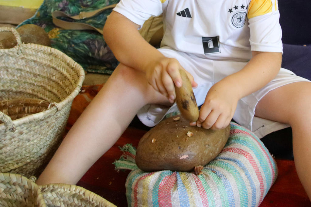 H&auml;nde und Beine einer sitzenden Person, die einen Holzst&ouml;&szlig;el auf eine gro&szlig;e braune Kartoffel auf einem gestreiften Kissen h&auml;lt; links zwei geflochtene K&ouml;rbe; Person tr&auml;gt wei&szlig;es Deutschland-Fu&szlig;balltrikot