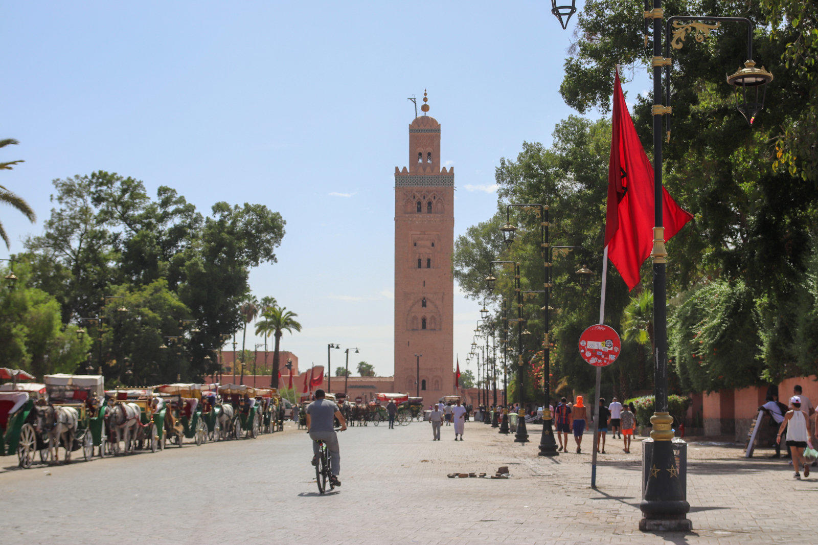 Breite Stra&szlig;e mit Fu&szlig;g&auml;ngern, einem Radfahrer und Pferdekutschen, im Hintergrund ein hoher r&ouml;tlicher Turm mit dekoriertem oberen Teil, rechts rote Fahnen und B&auml;ume, blauer Himmel
