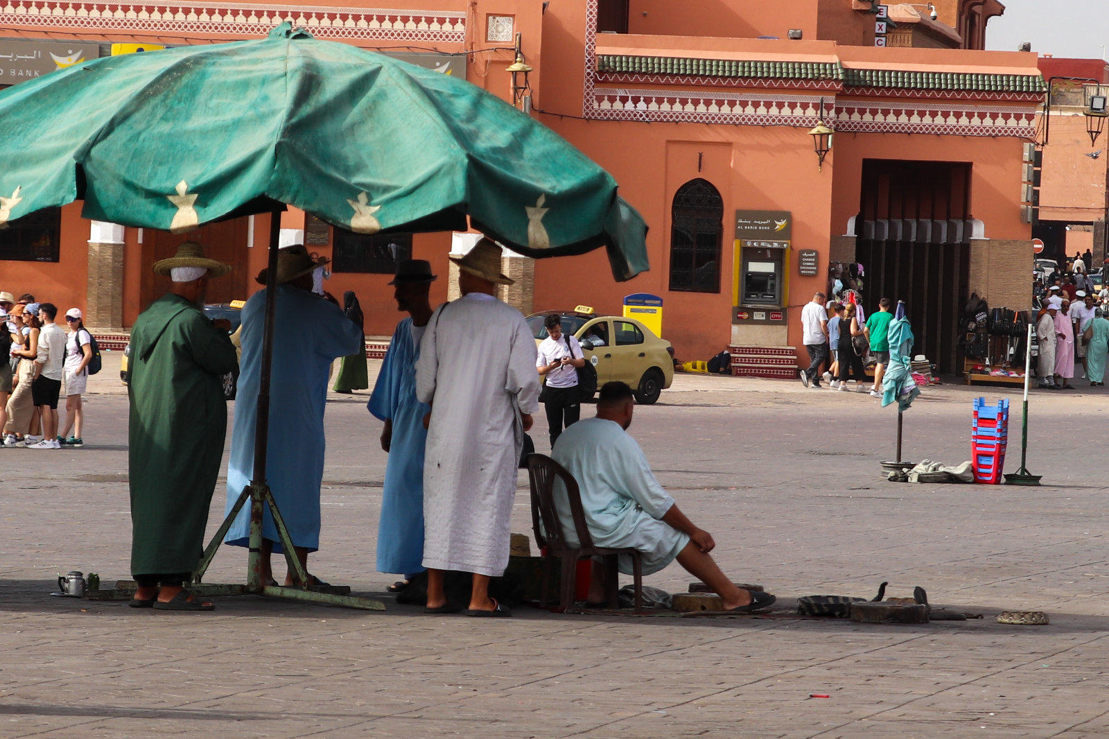 Gruppe von M&auml;nnern in langen Gew&auml;ndern unter einem gro&szlig;en gr&uuml;nen Sonnenschirm auf einem Platz; im Hintergrund orangefarbene Geb&auml;ude, ein geparkter kleiner gelber Wagen und weitere Personen