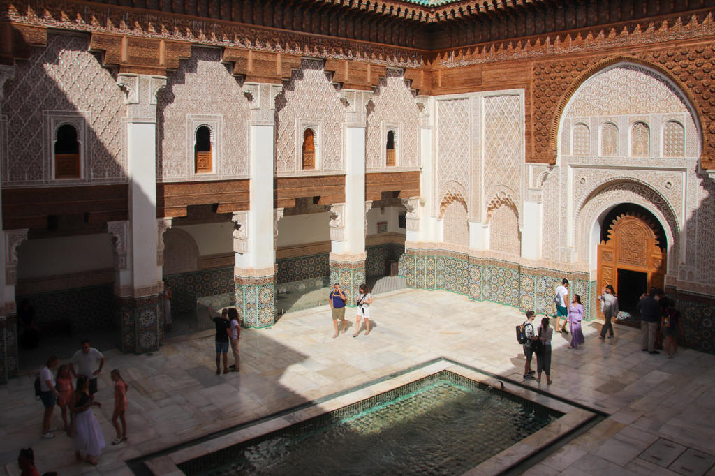 Innenhof mit rechteckigem Wasserbecken in der Mitte, hellen Bodenplatten, Reihe von S&auml;ulen mit verzierten B&ouml;gen, gemusterte Fliesen an den unteren W&auml;nden, geschnitzter Stuck an den oberen W&auml;nden, mehrere Personen stehend oder gehend, Licht- und Schat