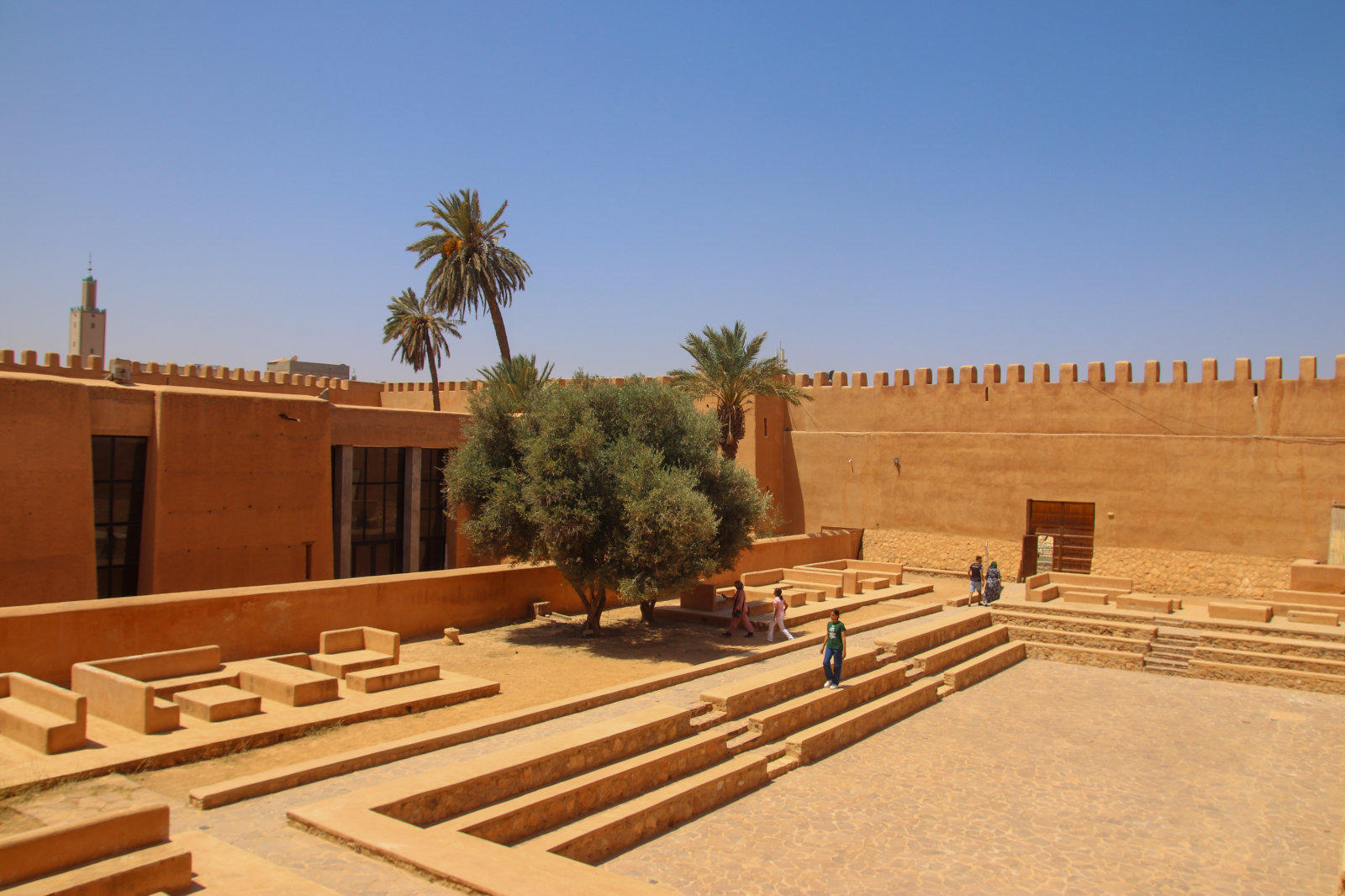 Innenhof mit sandfarbenen Terrassen und rechteckigen, abgestuften Becken; zentraler Baum, Palmen, hohe zinnenbekr&ouml;nte Mauer, blauer Himmel, einige Personen