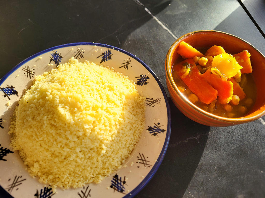 Teller mit H&uuml;gel aus gelben feinen K&ouml;rnern neben brauner Schale mit orangefarbenen Gem&uuml;sest&uuml;cken und gr&uuml;nen Erbsen auf dunklem Tisch