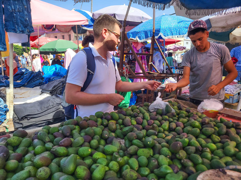 Haufen gr&uuml;ner Avocados im Vordergrund. Zwei M&auml;nner an einem Marktstand unter bunten Sonnenschirmen; links ein Mann mit wei&szlig;em Poloshirt und Rucksack &uuml;bergibt Geld, rechts ein Mann mit grauem T‑Shirt und Kappe nimmt es entgegen. Plastikbeutel und Waag