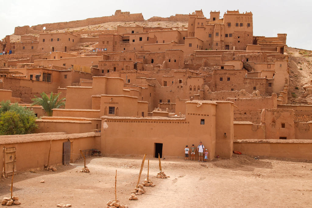 Mehrst&ouml;ckige, sandfarbene Lehmbauten an einem Hang; im Vordergrund ein offener Hof mit drei Personen neben einer Mauer und einigen Palmen