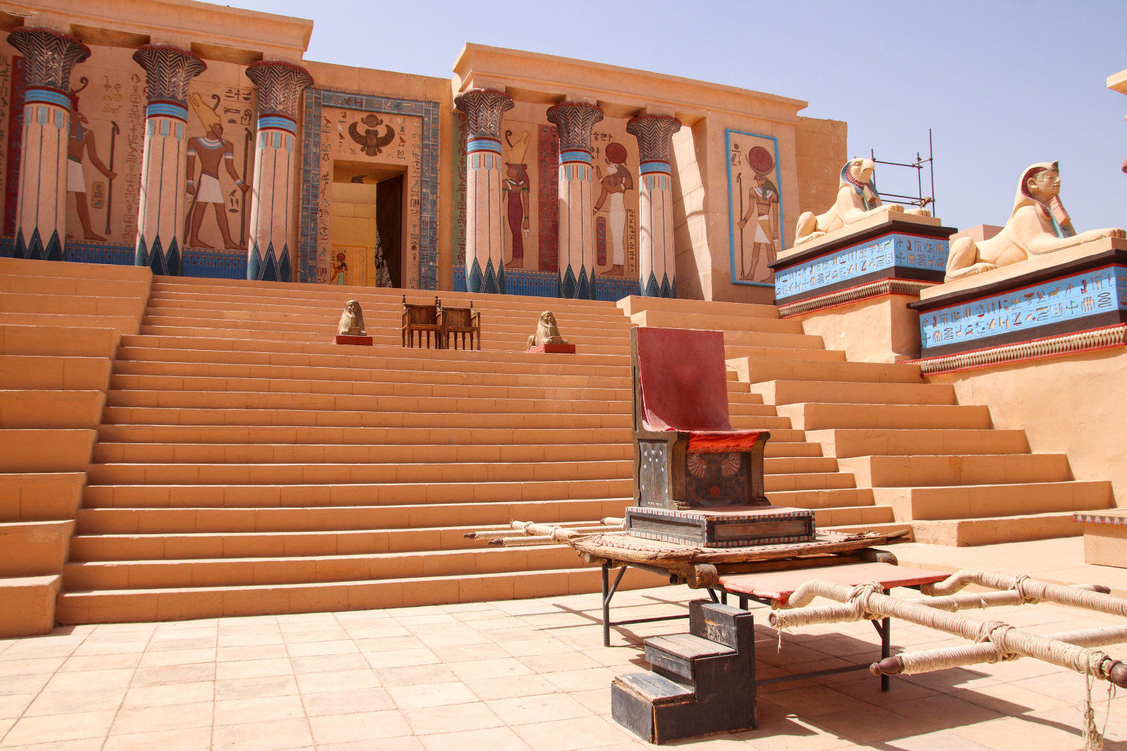 Breite steinerne Treppe vor einer Fassade mit bemalten S&auml;ulen und Wandmalereien; rechts zwei liegende L&ouml;wenstatuen; auf einem Podest ein roter Thron; davor Tisch, Kisten und Liegen auf gepflastertem Hof
