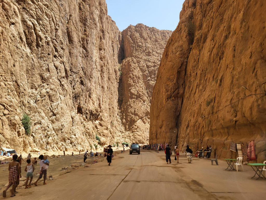 Schmale Felsenschlucht mit hohen, steilen Felsw&auml;nden, sandigem Boden, mehreren Personen, einem geparkten Fahrzeug und Tischen/St&auml;nden unter klarem Himmel