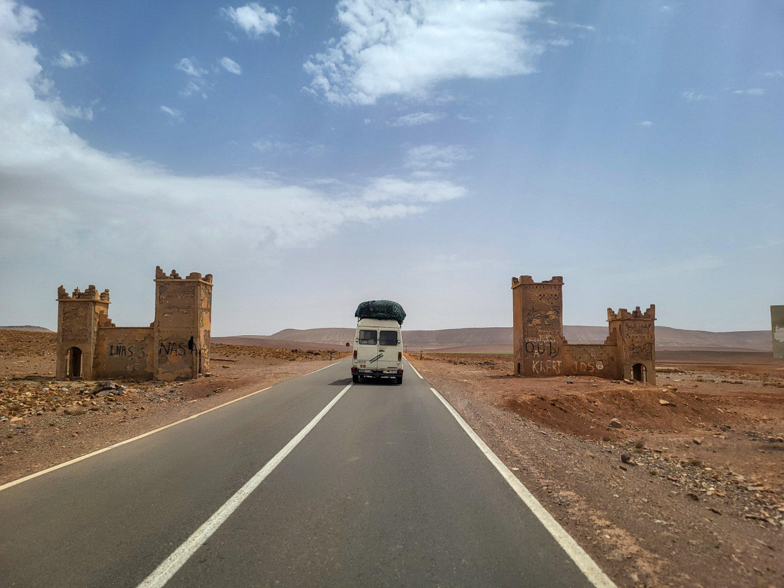 Asphaltierte gerade Stra&szlig;e in einer W&uuml;stenlandschaft, mittig ein wei&szlig;er Camper von hinten, sandfarbene Turmruinen links und rechts, teils bew&ouml;lkter Himmel