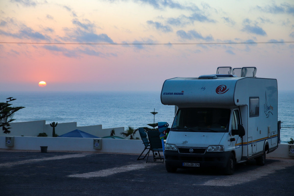 Wohnmobil auf gepflastertem Platz am Meer bei Sonnenuntergang; zwei Klappst&uuml;hle und kleiner Tisch daneben; niedrige Mauer und bew&ouml;lkter Himmel