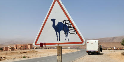 Dreieckiges Schild mit blauer Kamel-Silhouette und schwarzer Graffiti auf einem Metallpfosten am Stra&szlig;enrand; asphaltierte Stra&szlig;e, wei&szlig;es Fahrzeug f&auml;hrt weg, Geb&auml;ude, karges W&uuml;stenland und blauer Himmel