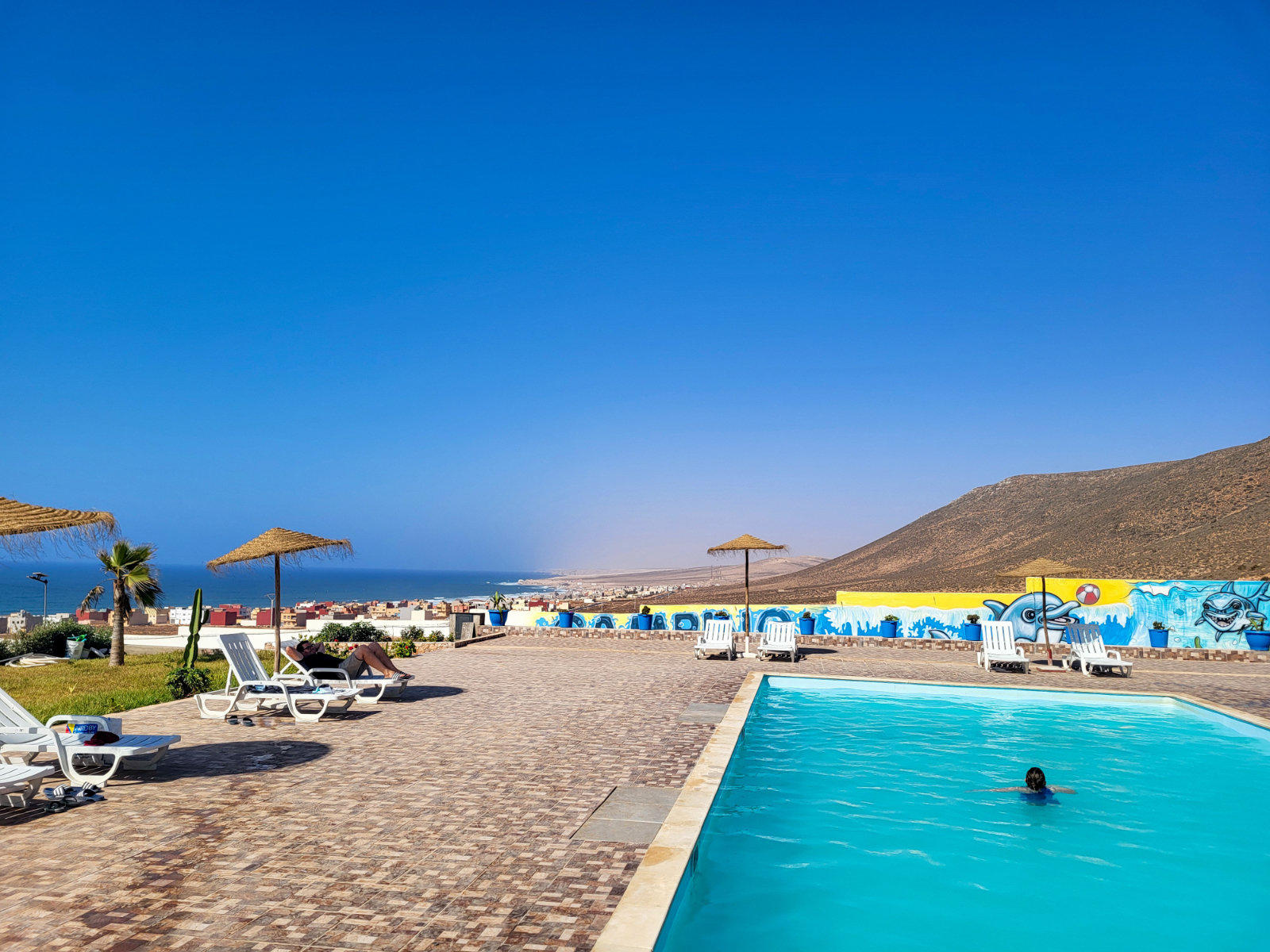 Freiluftpool mit einer Person im Wasser, Liegest&uuml;hle und Strohh&uuml;te auf gepflasterter Terrasse, Meer und H&uuml;gel im Hintergrund, blauer Himmel