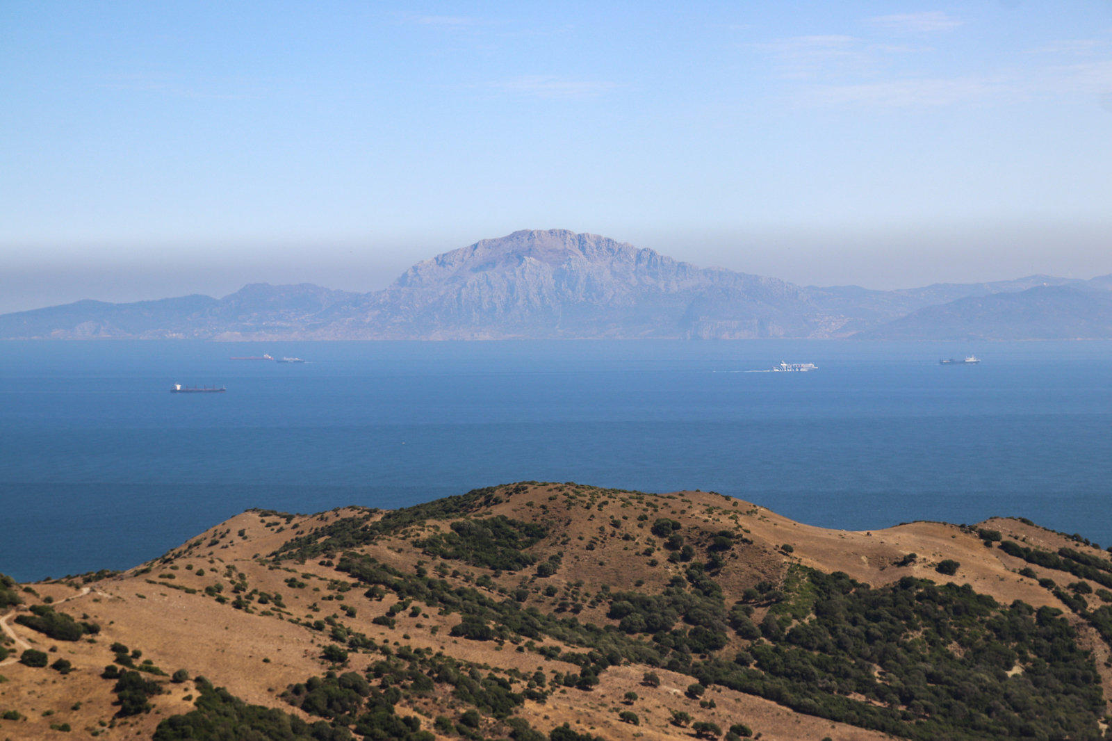 Vordergrund: trockene, grasbewachsene H&uuml;gel mit vereinzelten Str&auml;uchern; Mittelgrund: ruhiges Meer mit mehreren Schiffen; Hintergrund: entfernte Gebirgsformation &uuml;ber dem Meer unter klarem Himmel