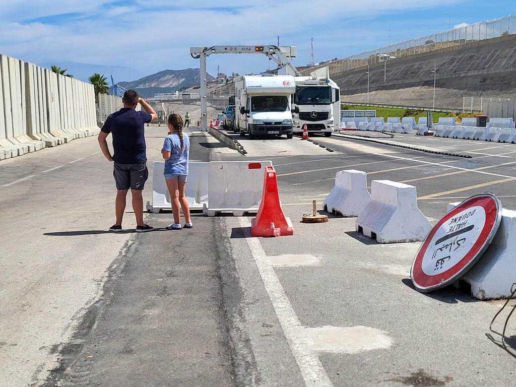 Zwei Personen stehen auf einer gesperrten Fahrspur neben Betonbarrieren; zwei wei&szlig;e Lastwagen weiter vorn; roter Verkehrsleitkegel und umgefallenes Verkehrsschild im Vordergrund; Berge und blauer Himmel im Hintergrund