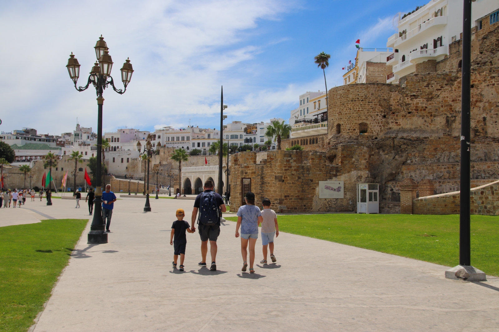 Promenade mit Personen vor einer steinernen Stadtmauer, Stra&szlig;enlaternen, Rasen und H&auml;usern unter blauem Himmel