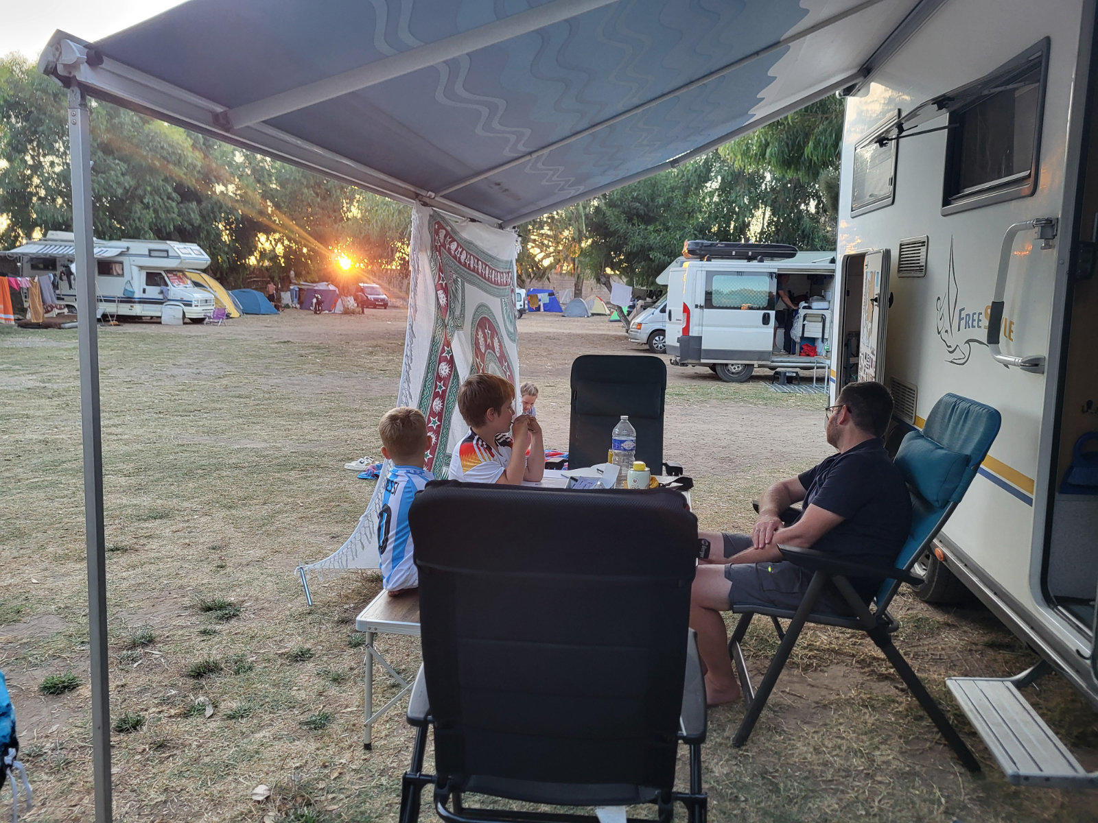 Drei Personen sitzen an einem Klapptisch unter der ausgefahrenen Markise eines Wohnmobils; Flaschen und Becher auf dem Tisch; im Hintergrund Zelte, Wohnwagen und die Sonne am Horizont