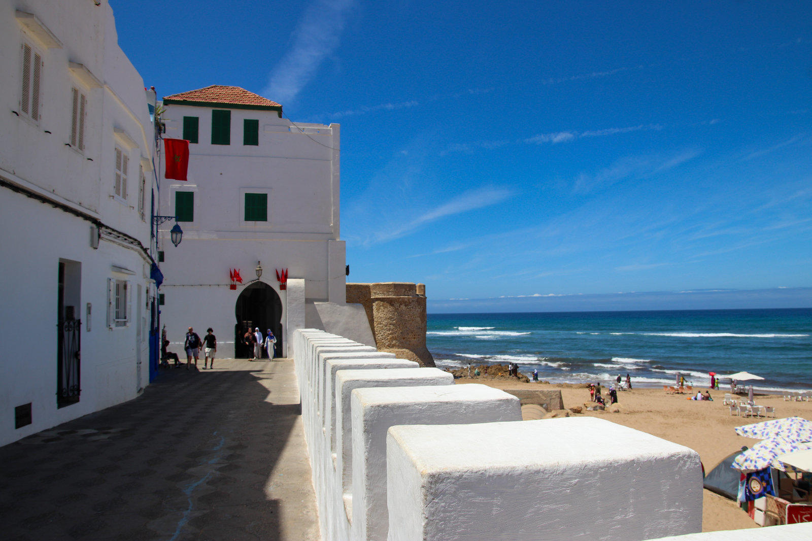Wei&szlig;e H&auml;user mit gr&uuml;nen Fensterl&auml;den an einer Promenade, Personen im Schatten, niedrige wei&szlig;e Br&uuml;stung, Sandstrand mit Sonnenschirmen, Meer und blauer Himmel