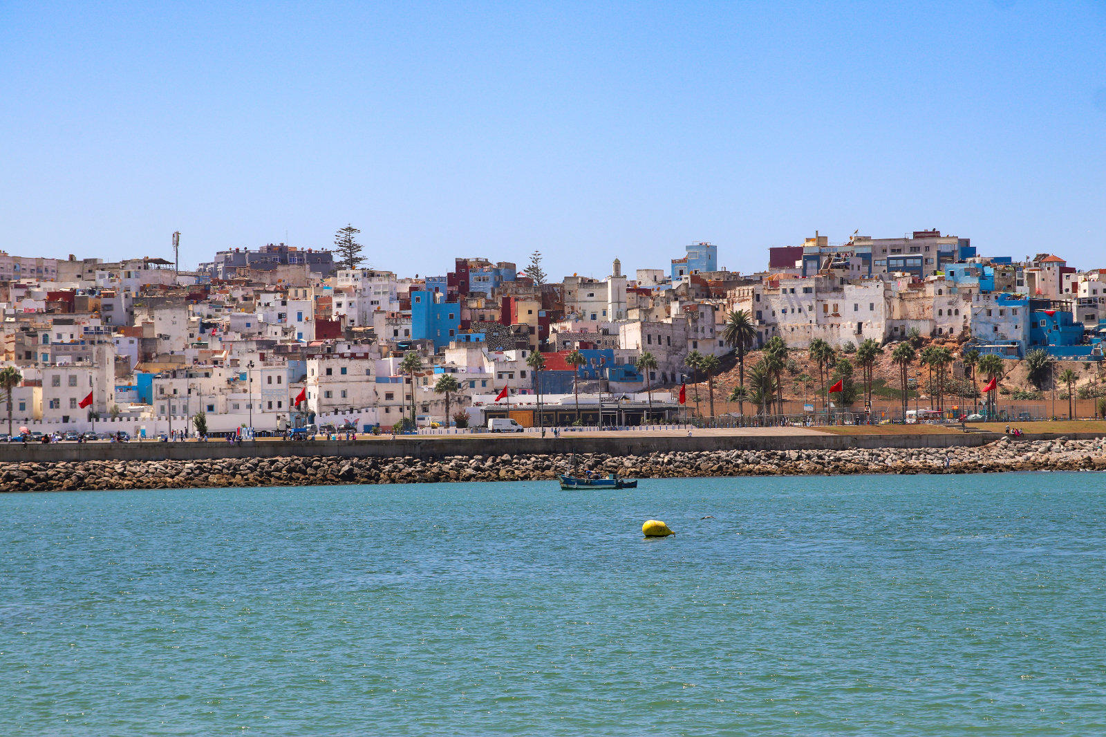 Blick auf dicht aneinander gebaute wei&szlig;e und farbige H&auml;user am Ufer, davor Wasser mit einer gelben Boje und blauem Himmel