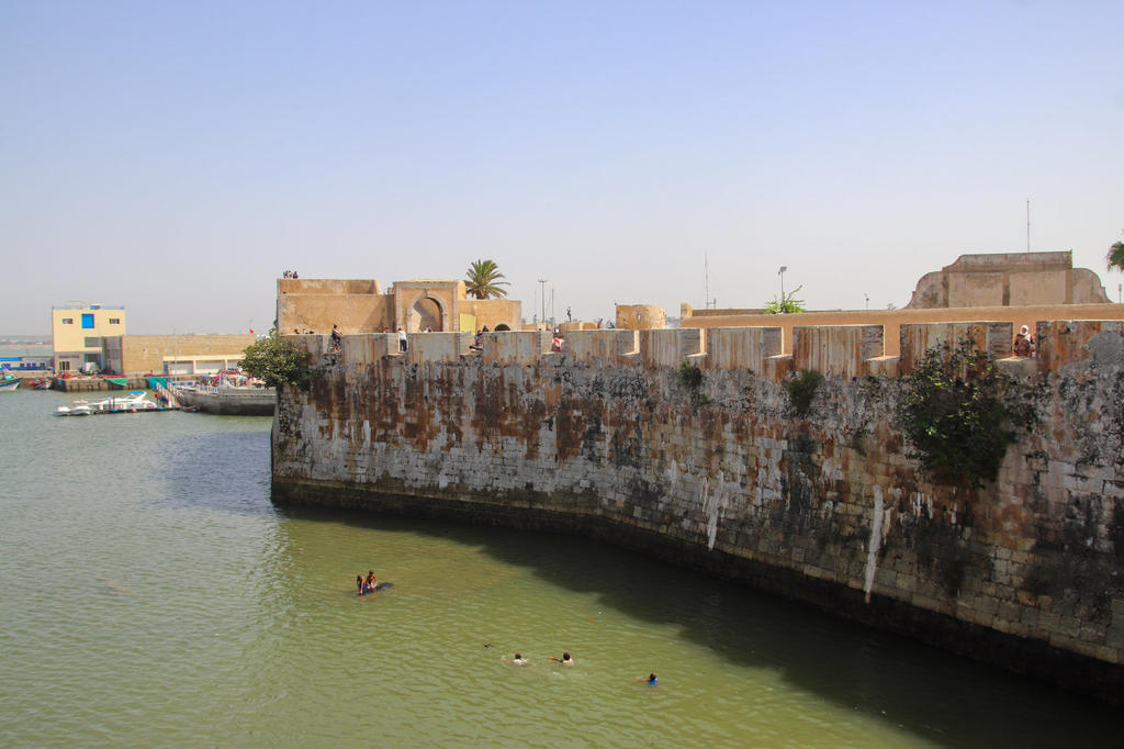 Steinerne Hafenmauer am Wasser; mehrere Personen schwimmen nahe der Mauer; Boote und niedrige Geb&auml;ude im Hintergrund; klarer Himmel