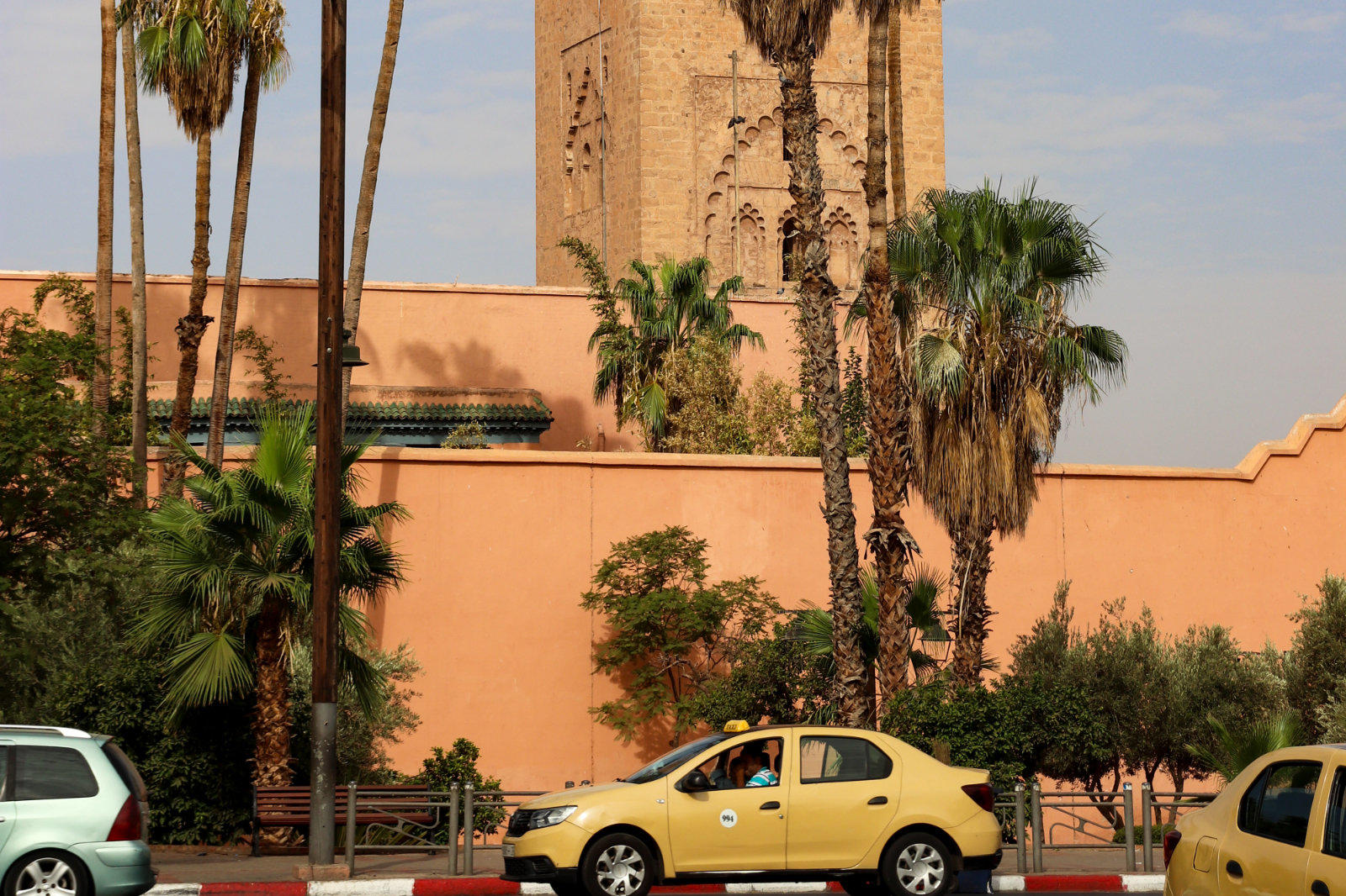 Stra&szlig;e mit Autos am Rand vor einer rosa-beigen Mauer, hohen Palmen und einem hohen, verzierten Turm im Hintergrund