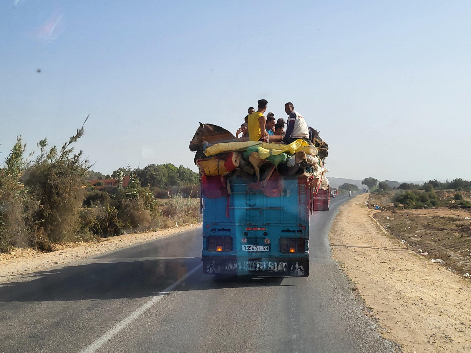 R&uuml;ckansicht eines blauen Lastwagens auf einer Landstra&szlig;e, mehrere Personen sitzen auf bunter Ladung unter klarem Himmel