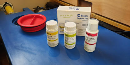 Drei kleine wei&szlig;e Flaschen mit Etiketten in Gelb, Gr&uuml;n und Rot vor einer wei&szlig;en Schachtel auf einer blauen Unterlage; links eine rote Sch&uuml;ssel; Holzpaneel im Hintergrund