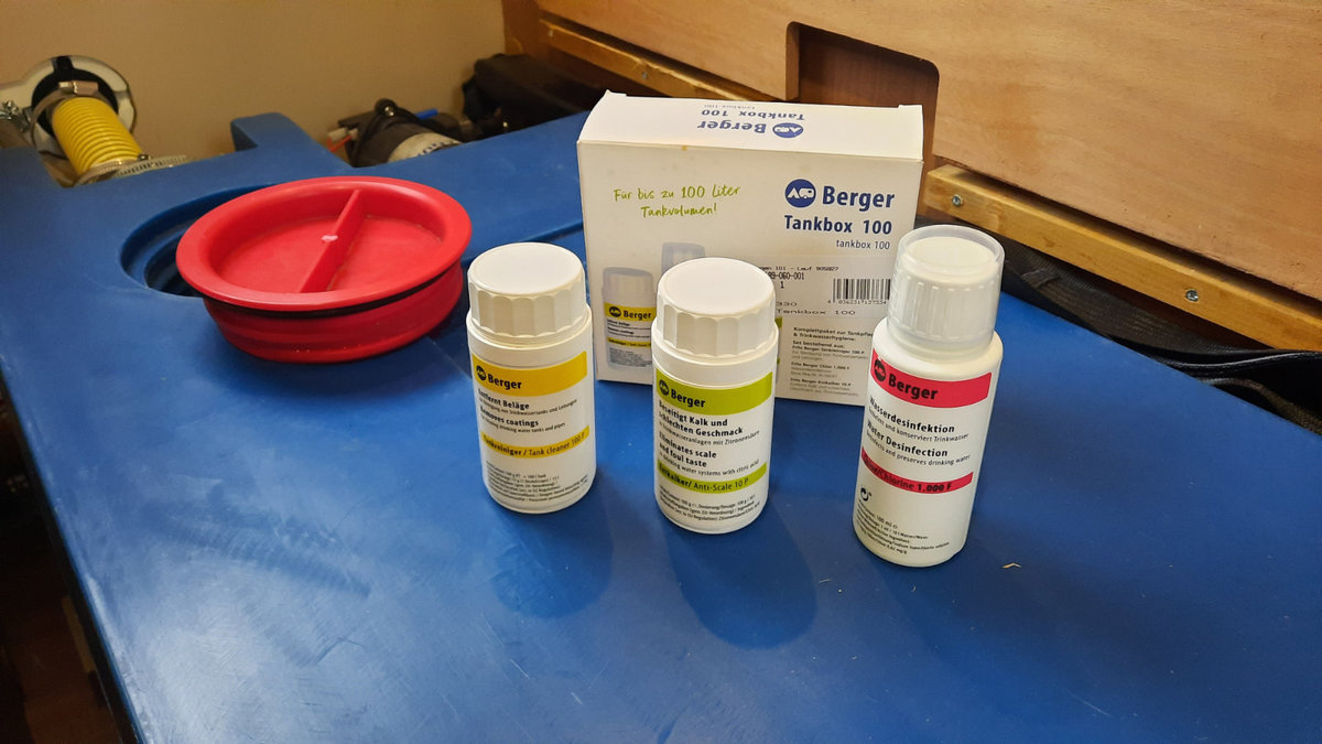 Drei kleine wei&szlig;e Flaschen mit Etiketten in Gelb, Gr&uuml;n und Rot vor einer wei&szlig;en Schachtel auf einer blauen Unterlage; links eine rote Sch&uuml;ssel; Holzpaneel im Hintergrund