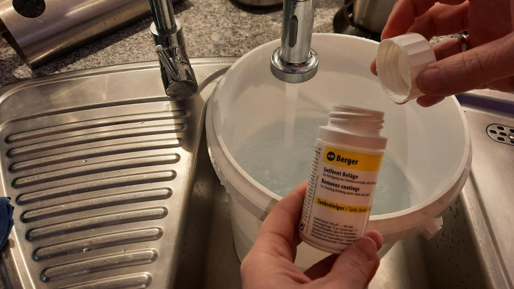 Zwei H&auml;nde halten eine gelbe Tablettenflasche und einen wei&szlig;en Deckel mit einer Tablette &uuml;ber einem wei&szlig;en Eimer unter einem laufenden K&uuml;chenwasserhahn