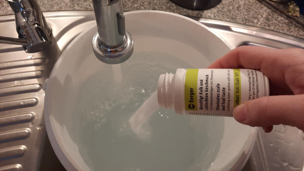 Hand h&auml;lt eine wei&szlig;e Flasche und kippt wei&szlig;es Pulver in eine mit Wasser gef&uuml;llte Plastiksch&uuml;ssel; Sp&uuml;le und Wasserhahn sichtbar