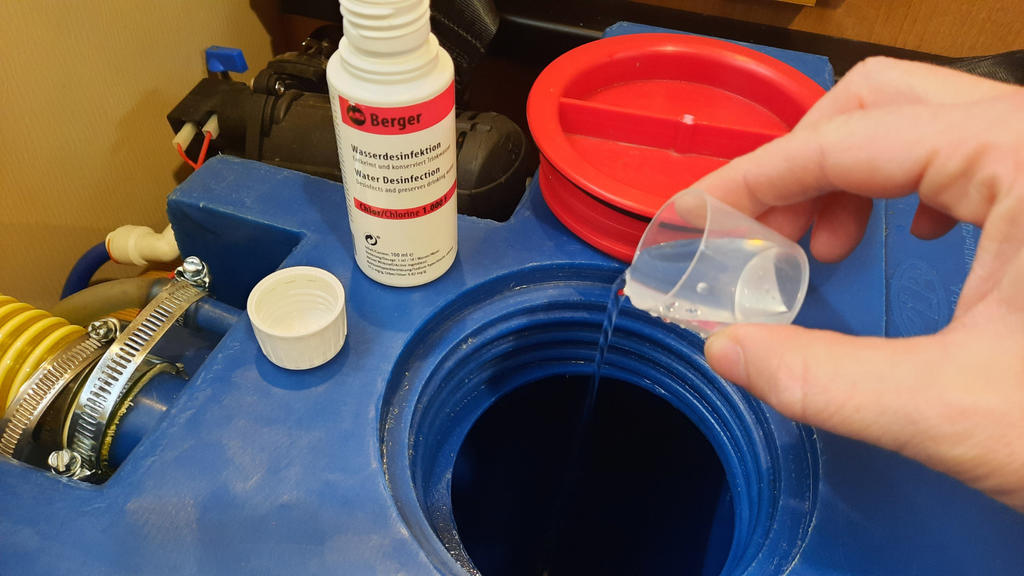 Hand gie&szlig;t Fl&uuml;ssigkeit aus kleinem transparenten Messbecher in offene blaue Kunststoff&ouml;ffnung; wei&szlig;e Flasche und rote Schale daneben sichtbar