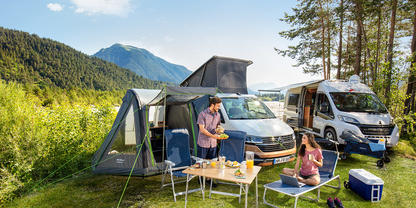 Zwei Personen neben zwei Wohnmobilen auf einer Wiese; Campingtisch mit Geschirr, K&uuml;hlbox, Campingst&uuml;hle, Vorzelt am linken Wohnmobil; B&auml;ume, Berge und blauer Himmel im Hintergrund
