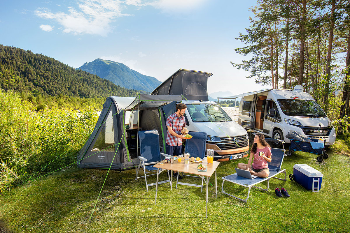 Zwei Personen neben zwei Wohnmobilen auf einer Wiese; Campingtisch mit Geschirr, K&uuml;hlbox, Campingst&uuml;hle, Vorzelt am linken Wohnmobil; B&auml;ume, Berge und blauer Himmel im Hintergrund