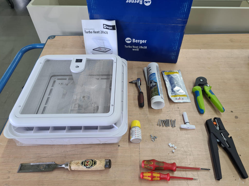 Werkzeuge und Verpackungen auf einem Holztisch: wei&szlig;er Kunststoffrahmen mit transparentem Einsatz, zwei Kartons (blau, wei&szlig;), Kartusche, kleine gelbe Flasche, zwei rote Schraubendreher, Holzspachtel mit Metallklinge, mehrere kleine Metallteile und ve