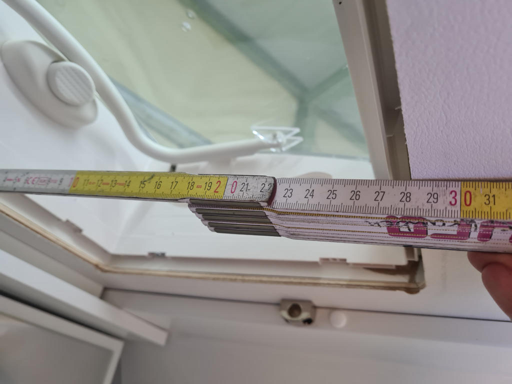 Hand h&auml;lt ein ausgeklapptes Ma&szlig;band &uuml;ber einen wei&szlig;en Fensterrahmen; sichtbare Skala mit Zahlen, Gelenk, Fenstergriff und Fensterglas