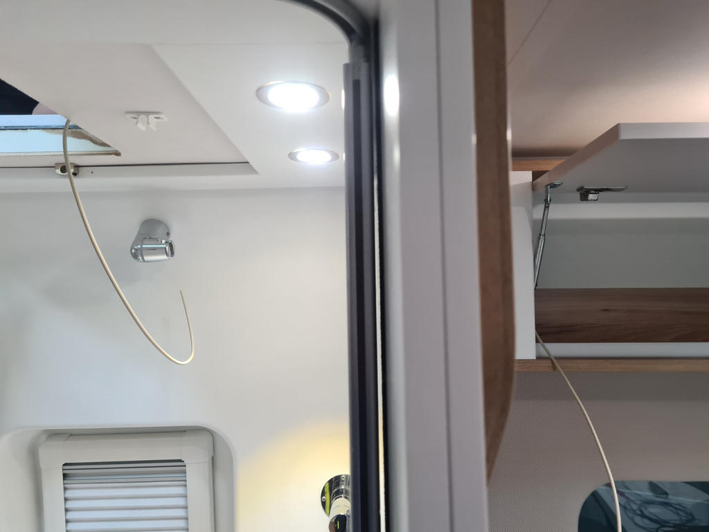 Wei&szlig;e Decke und Wand mit zwei runden Deckenleuchten, rechteckige Dachluke oben links, Metallhaken und h&auml;ngendes gelbes Kabel, geschlossenes Fenster mit Jalousie unten links, offenes Holzfach mit Scharnieren rechts, schwarzer vertikaler Rahmen in der