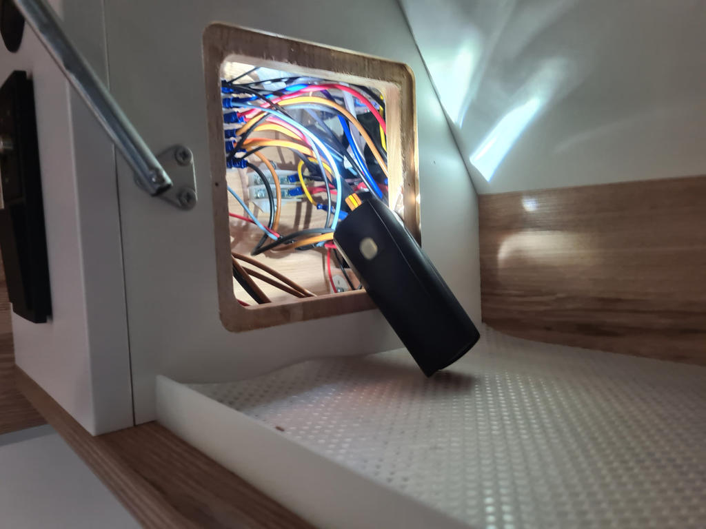 Rechteckige &Ouml;ffnung in einer Holzverkleidung mit sichtbaren bunten Kabeln, einer schwarzen Taschenlampe an der Kante, einem Metalllampenarm und einer hellen, strukturierten Fl&auml;che